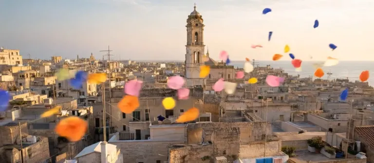 Spot MYRA realizzato con AI generativa: petali colorati sopra i tetti di Bari Vecchia, con il campanile e il mare sullo sfondo